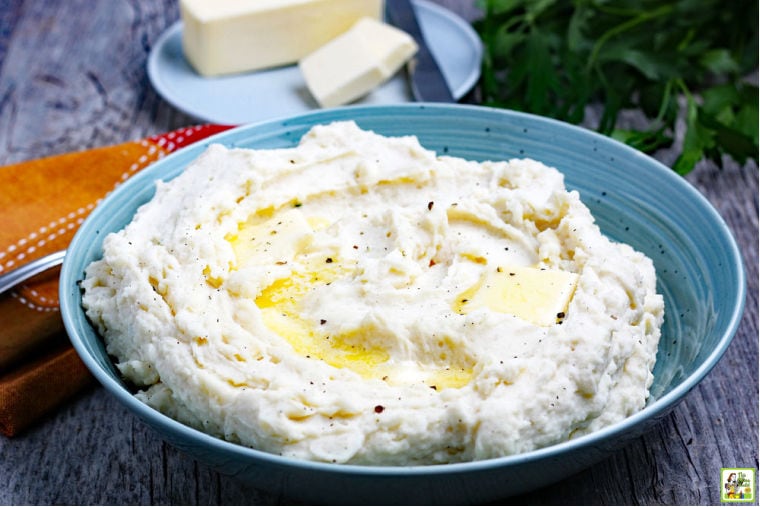 A blue bowl of Crockpot Mashed Potatoes with multicolored napkin and a plate with a stick of butter and a knife in the background.