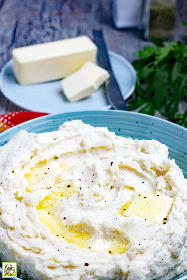 Buttery mashed potatoes in a blue bowl with a stick of butter on a blue plate in the background.