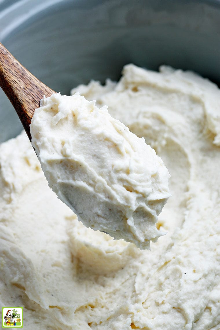 Closeup of a spoon of mashed potatoes above a slow cooker of mashed potatoes.