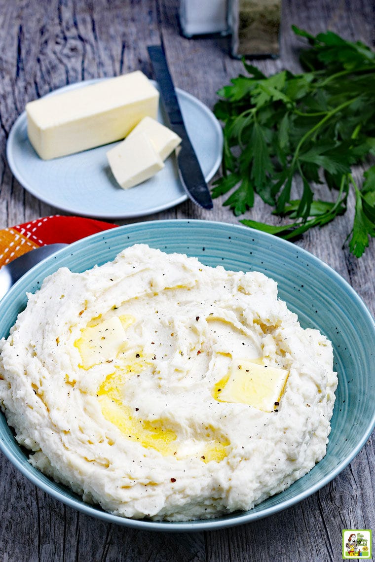 A blue plate of buttery mashed potatoes with more butter in the background on a blue plate with a knife.