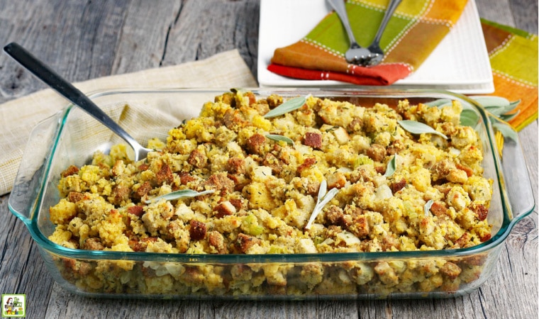 A glass casserole dish with Gluten Free Cornbread Stuffing, sage leaves, serving spoon with napkins, plates and forks in the background.