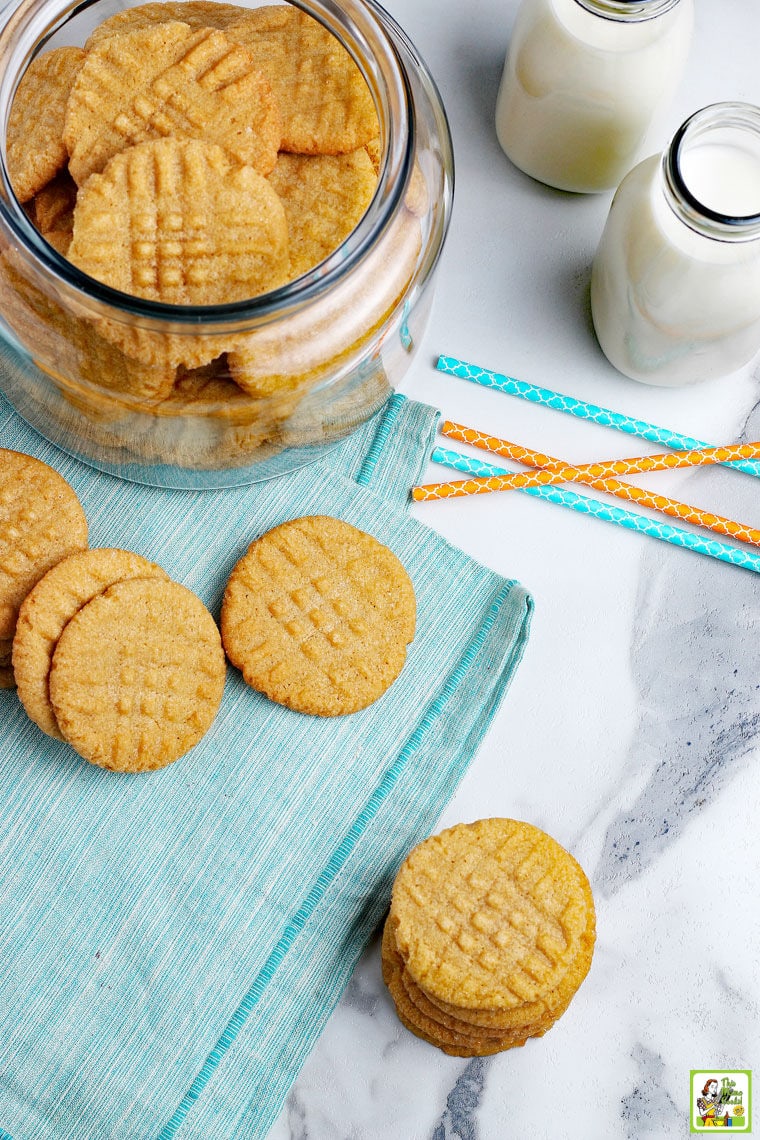Overhead shot of peanut butter cookies on a blue napkin with a jar of cookies , blue and orange paper straws, and milk bottles.