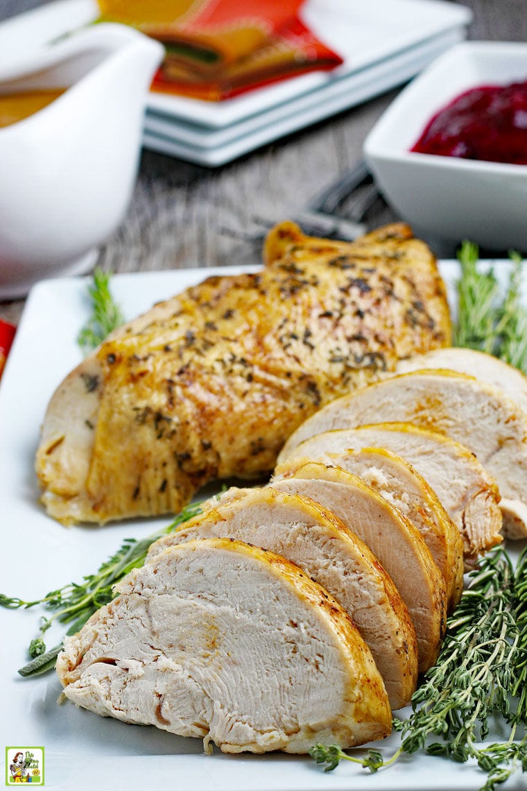 Cooked and sliced roasted turkey breast on a white platter with herbs with gravy, cranberry sauce, plates, and napkins.