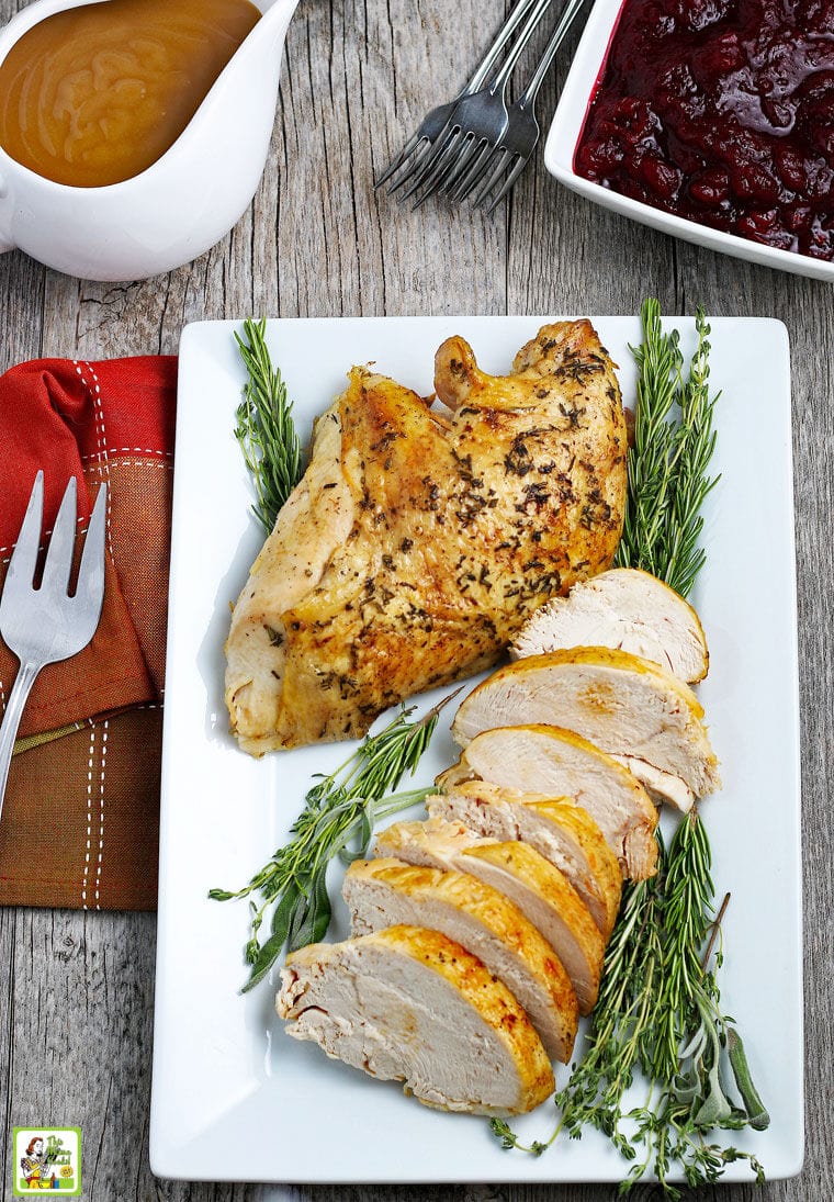 Cooked and sliced Turkey Breast on a white platter with herbs, napkin, serving fork, gravy boat, and bowl of cranberry sauce.