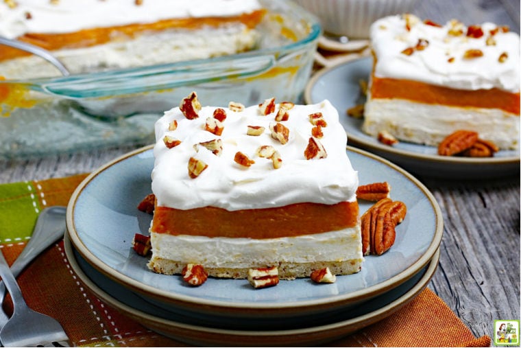 Blue plates of Pumpkin Cheesecake Lasagna with pecans on a multicolored placemat with forks with a baking dish in the background.