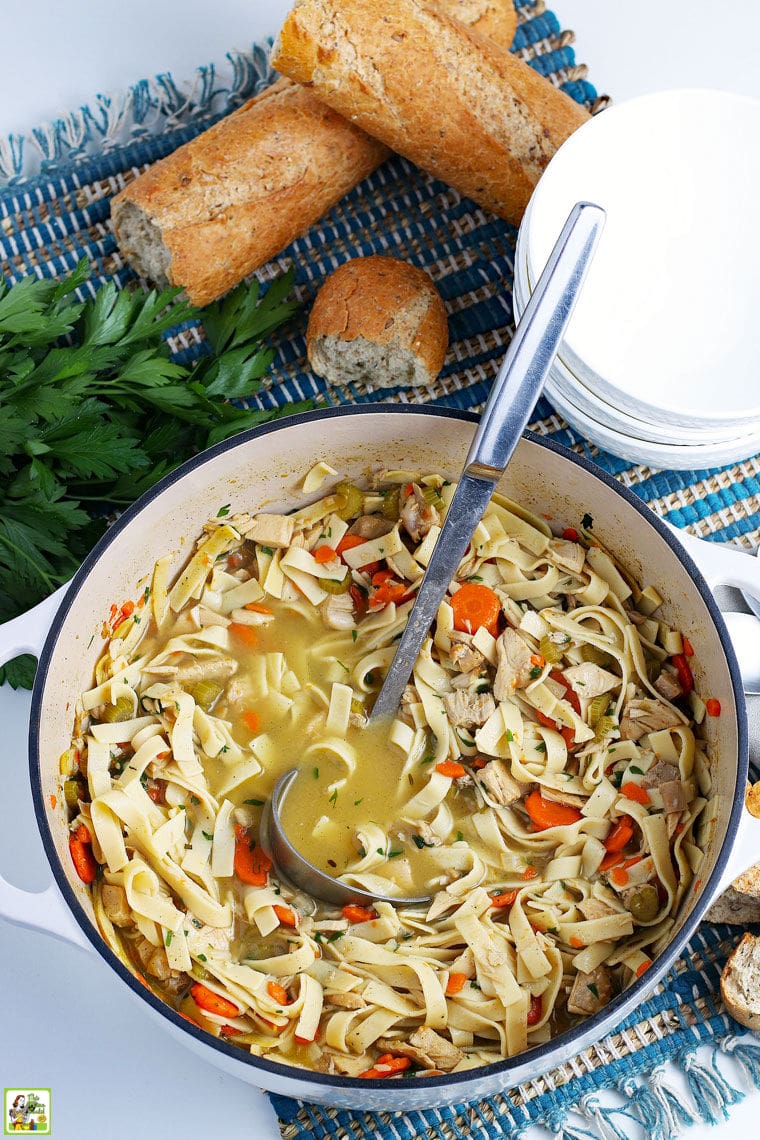 Overhead shot of a pot of turkey noodle soup with a ladle, soup bowls, and French bread, on a blue placemat.