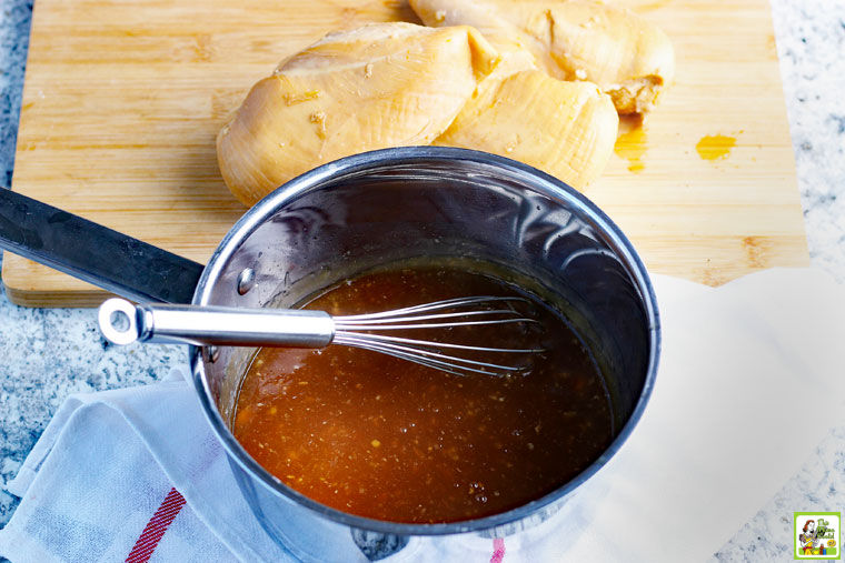 Cooking teriyaki sauce in saucepan with cooked chicken breasts in the background on a wooden cutting board.