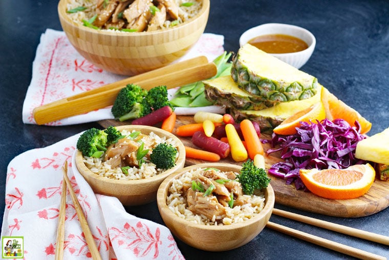 Wooden bowls of slow cooker teriyaki chicken on rice with broccoli with napkins, chopsticks, fruit, and bowls of sauce.