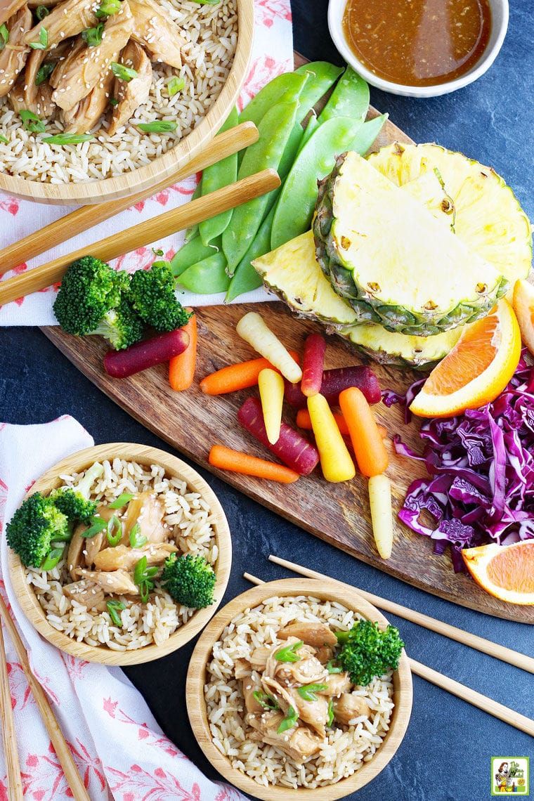 Bowls of Crock Pot Teriyaki Chicken on rice with vegetables on a wooden cutting board with serving utensils.