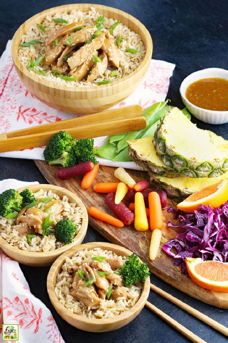 Teriyaki chicken with rice and broccoli in bowls with wooden serving utensils, vegetables, and fruits, colorful napkins, and a bowl of teriyaki sauce.