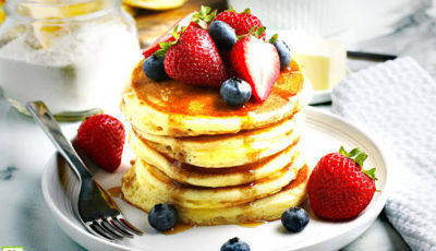 A stack of gluten free pancakes covered in syrup with fruit on a white plate.