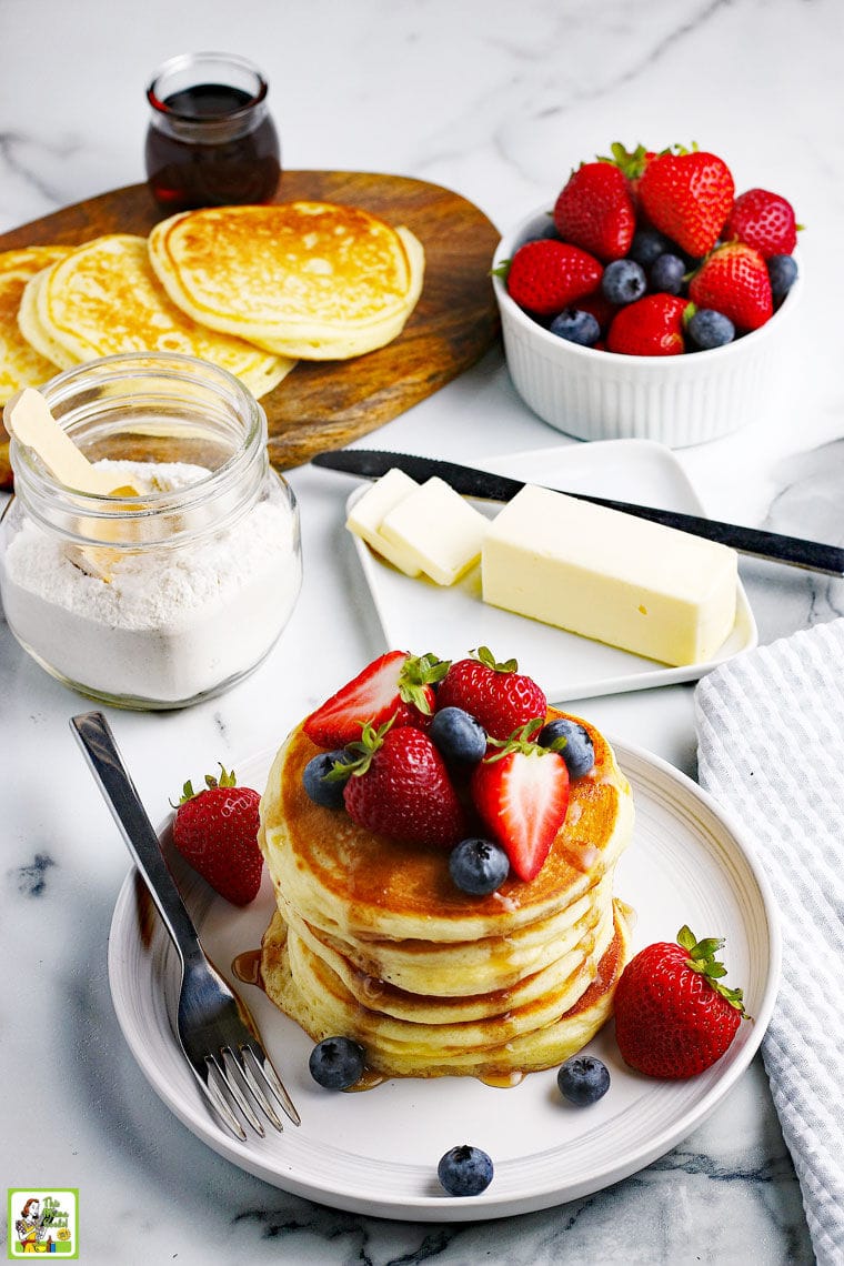 A stack of fluffy pancakes with fruit on a white plate with pancake mix in a glass jar, a stick of butter, more pancakes, and a bowl of fruit in the background.