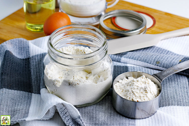 A glass jar and metal spoon filled with pancake mix on a gray and white tea towel with a lid and foodstuffs in the background.