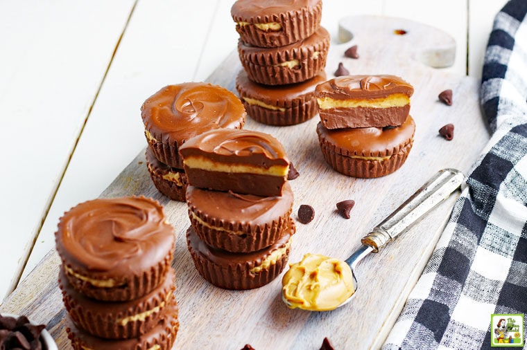 Stacks of Homemade Peanut Butter Cups on a wooden board with a tea towel, chocolate chips, and a spoon of peanut butter.