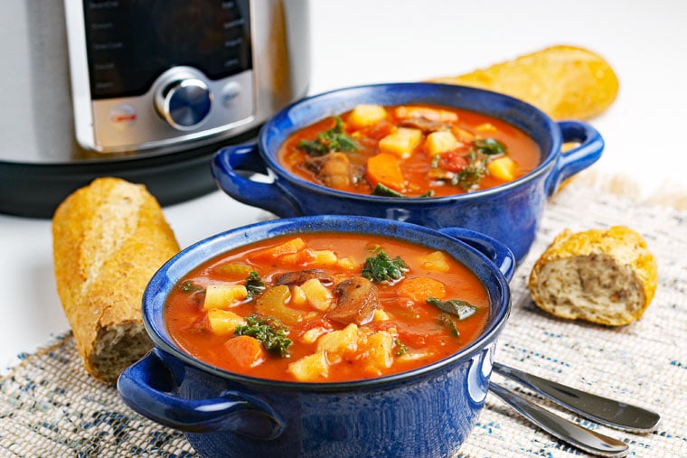 Two blue bowls of Instant Pot Vegetable Soup with crunchy French bread and spoons on a knit placemat with a pressure cooker in the background.