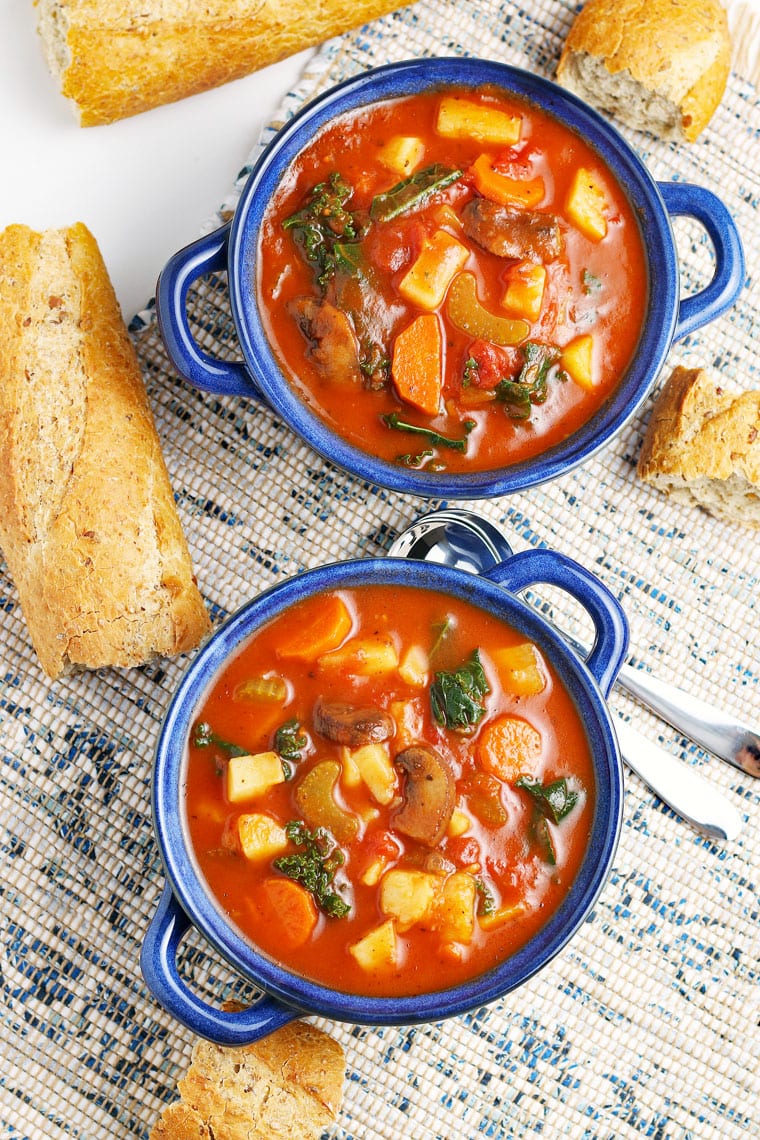 Two blue bowls of vegetable soup with French bread on blue and white placemats.
