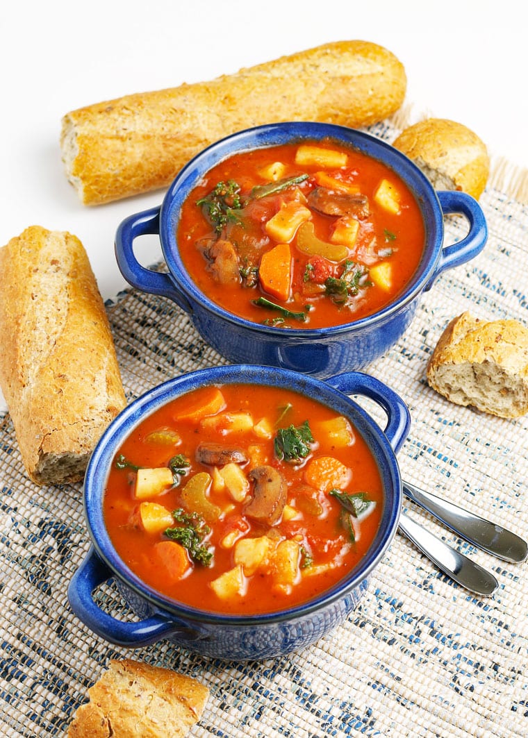 Two lug blue bowls of vegetable soup with French bread and spoons.