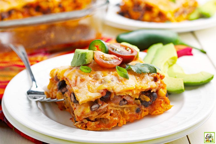 A plate of Chicken Enchilada Casserole with slices of avocados and a fork with a casserole and more plates in the background.