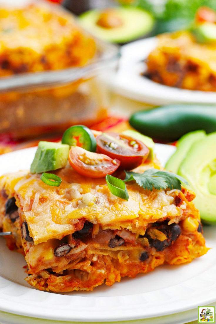 Close up of a slice of chicken enchilada casserole on a white plate with avocado and tomatoes.