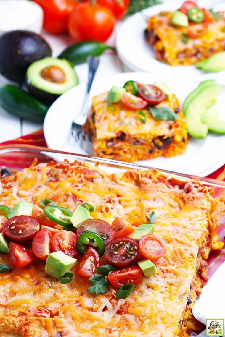 A glass casserole dish of enchiladas with avocados, tomatoes, plates of chicken enchiladas, and festive striped napkins. 