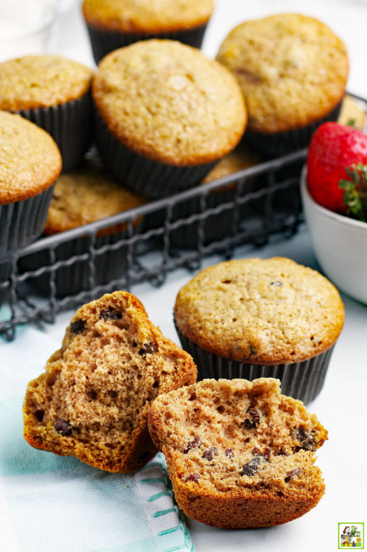 A wire basket of applesauce muffins with a broken muffin in front and a bowl of fresh strawberries with a blue and white tea towel.