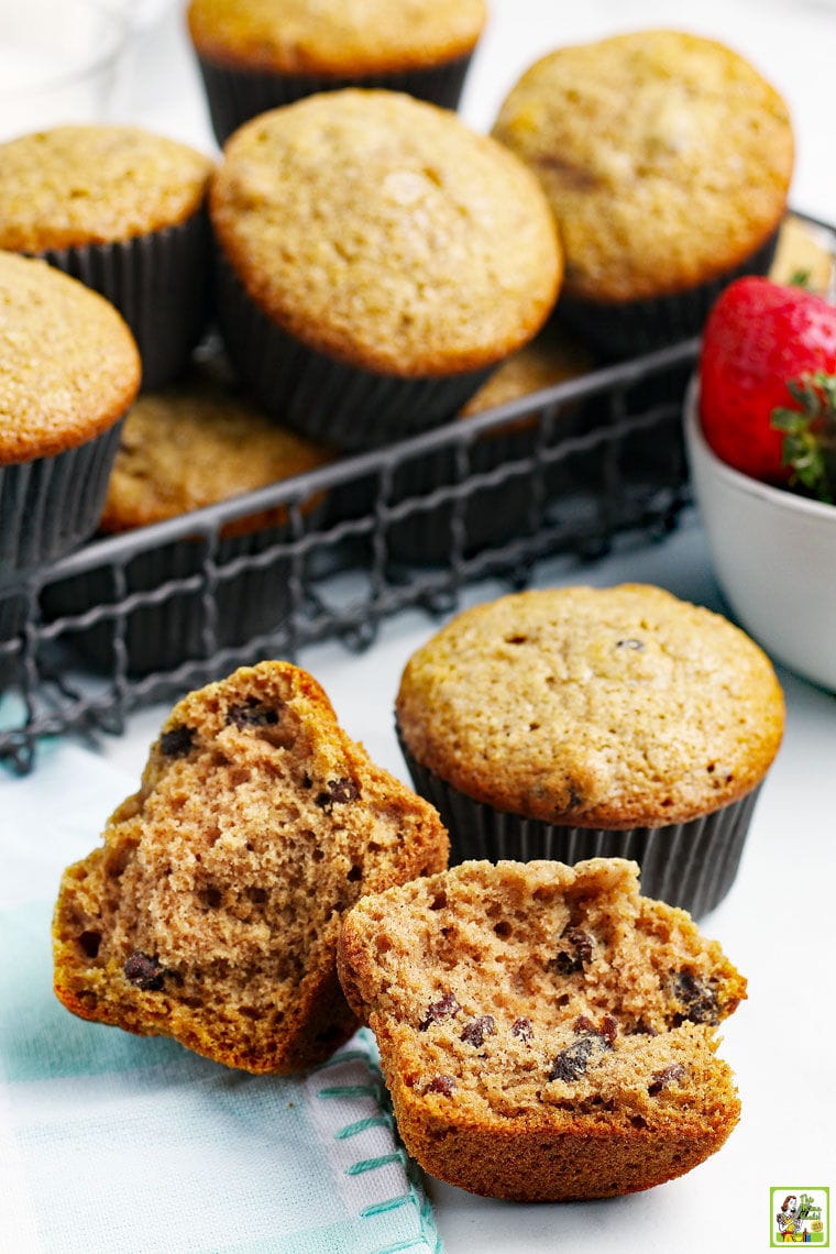 A wire basket of applesauce muffins with a broken muffin in front and a bowl of fresh strawberries with a blue and white tea towel.