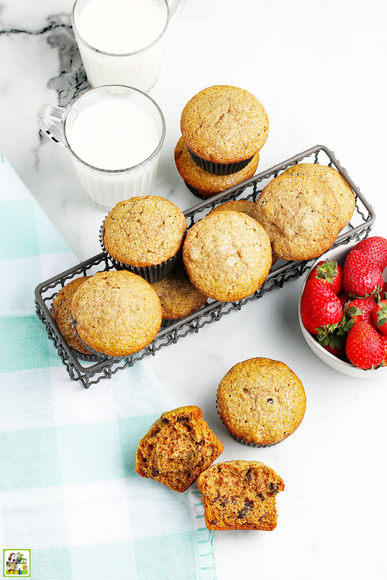 Muffins in a wire basket, two glasses of milk, a bowl of strawberries, and a blue and white napkin on a white countertop.