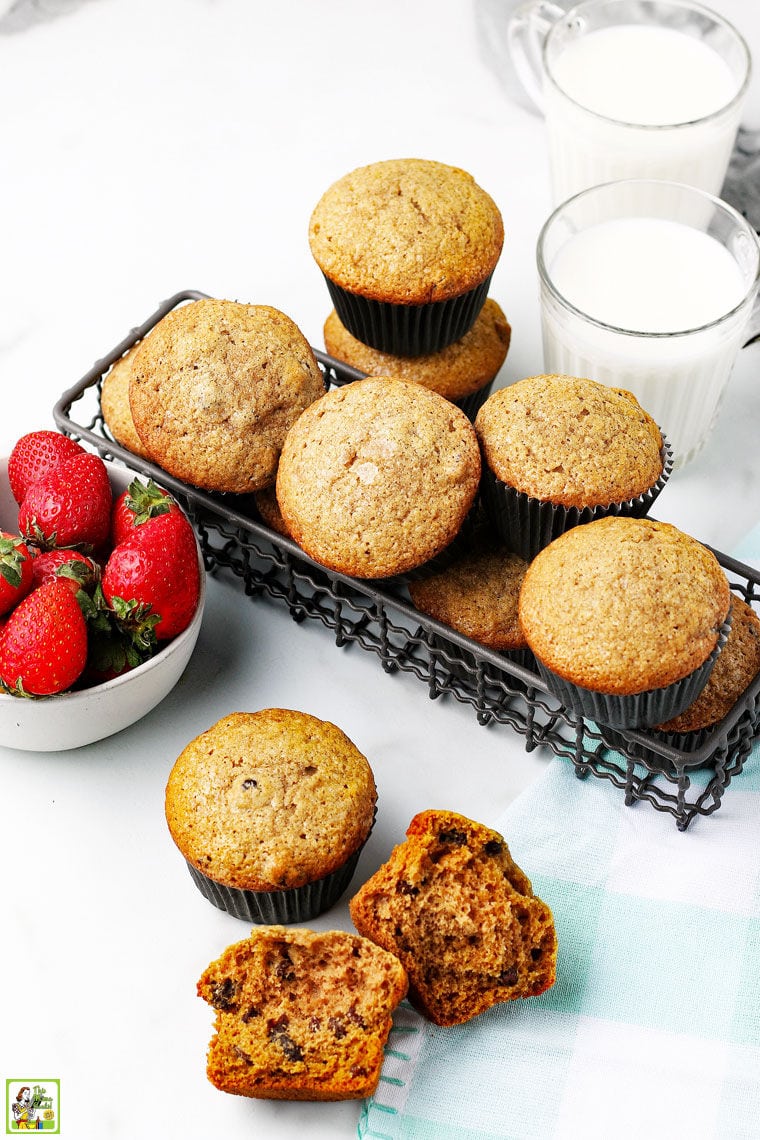 A bowl of strawberries, a basket of freshly baked muffins, a glass of milk, and a napkin on a white tabletop.