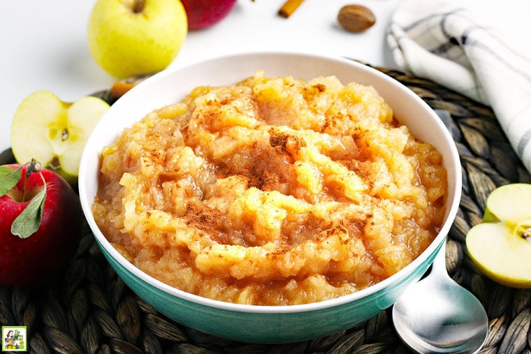 A bowl of Homemade Applesauce on a woven mat with a spoon and apples.