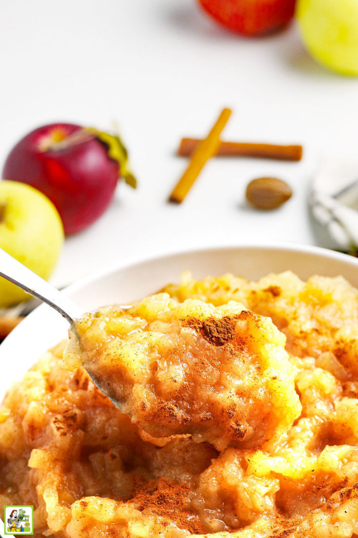 A spoon of homemade applesauce with cinnamon over a bowl of applesauce with apples, nutmeg, and cinnamon sticks in the background.