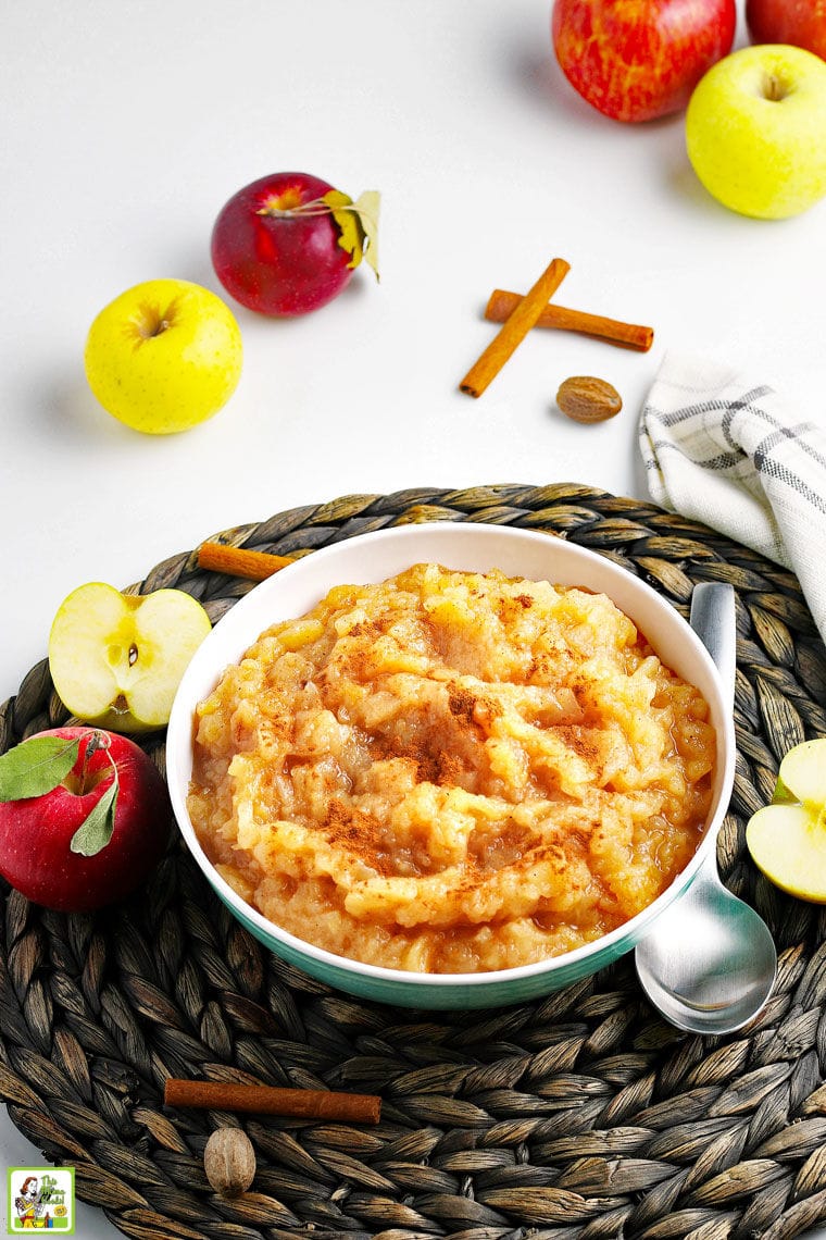 A bowl of applesauce with cinnamon with a serving spoon on a straw mat with apples, a tea towel, cinnamon sticks, and whole nutmeg.