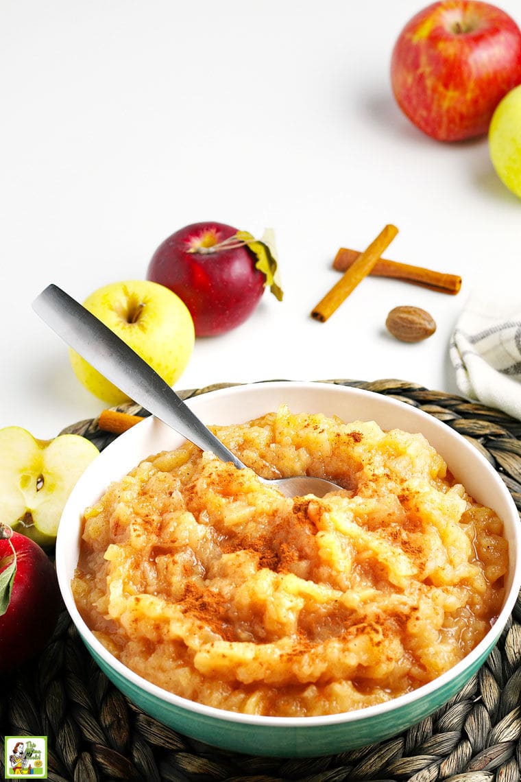 Closeup of a bowl of homemade apple with a serving sppon with apples, cinnamon, and nutmeg and a dishtowel in the background.