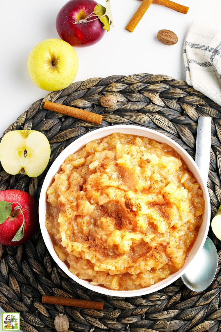 Overhead shot of a bowl of homemade apple with a serving spoon with apples, cinnamon, and nutmeg and a dishtowel on a woven mat.