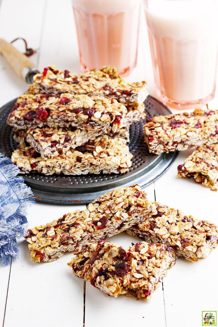 Stacks of homemade granola bars, a metal trivet, pink glasses of milk, a blue and white napkin, on a white wooden board.