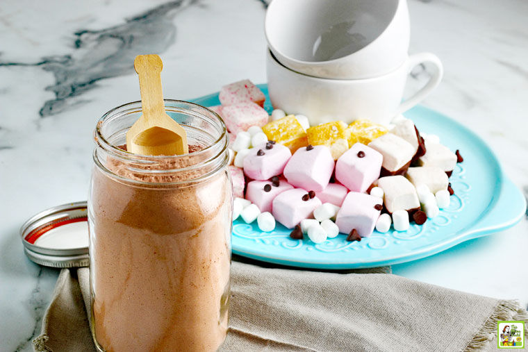 A mason jar of Homemade Hot Chocolate Mix, napkins, mugs, and a plate of hot cocoa toppings like marshmalows and chocolate chips.
