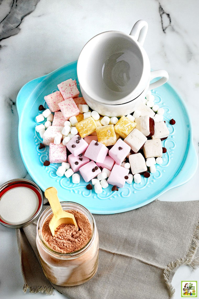 An overhead shot of a mason jar of hot chocolate mix with a wooden scoop and lid next to a blue plate of marshmallows, chocolate chips, and white ceramic mugs.