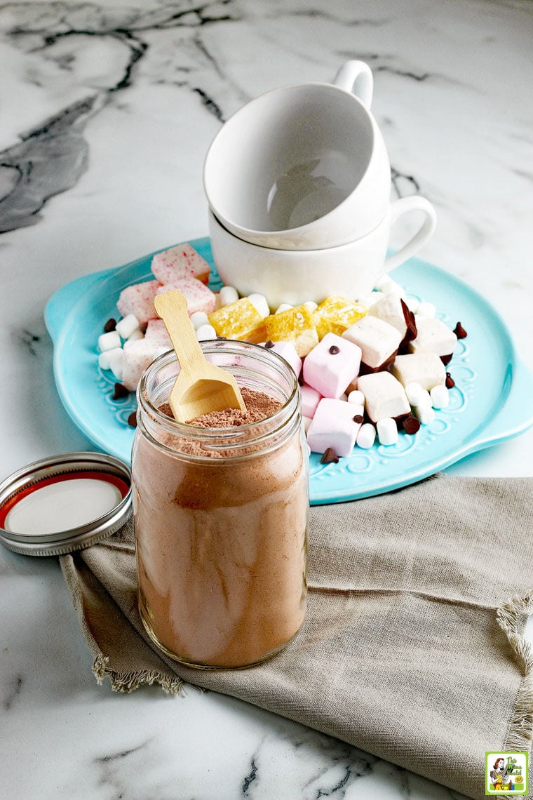 A large glass jar of hot chocolate mix with a wooden scoop and lid next to a blue plate of marshmallows, chocolate chips, and white ceramic mugs. on a marble table top with a gray napkin.