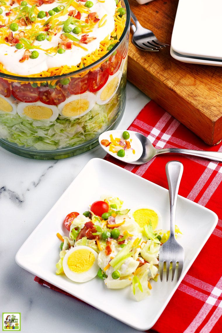 A plate of salad with fork and red napkin with a large glass bowl of layered salad in the background.