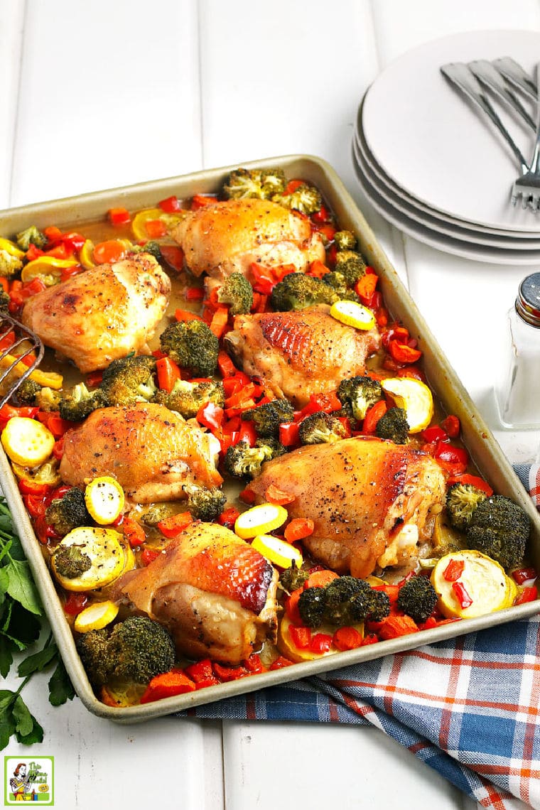 A roasting pan of cooked chicken thighs and vegetables with a colorful blue, orange and white napkin, white plates, and salt and pepper shakers.