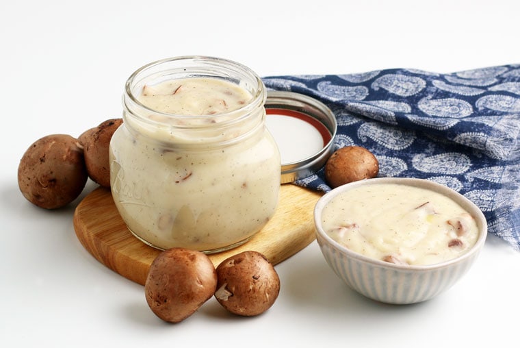 A small jar and small bowl of gluten free cream of mushroom soup on a wooden board with mushrooms and a blue napkin.