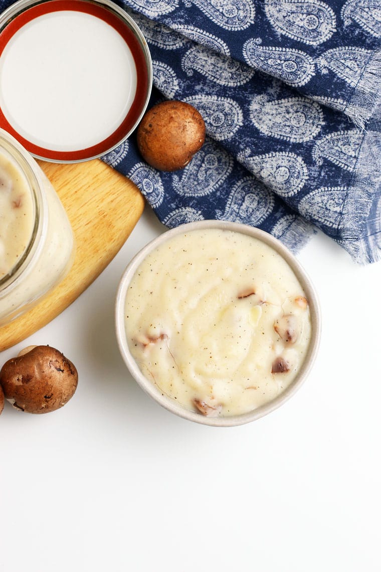 Overhead shot of a bowl and a mason jar of cream of mushroom soup on a wooden cutting board with mushrooms, jar lid, and a blue napkin.