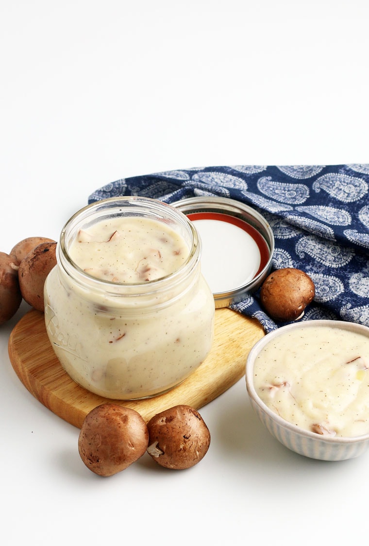Bowl and a glass jar of cream of mushroom soup on a wooden cutting board with mushrooms, jar lid, and a blue napkin.