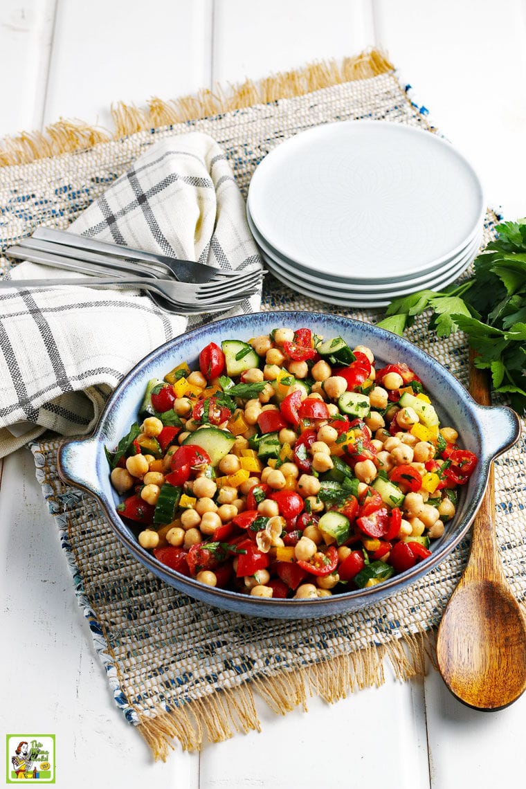 A bowl of a vegan garbanzo bean, tomatoes, cucumber, and bell pepper salad in a blue bowl with a wooden spoon, white plates, forks, napkins, herbs, on a rustic placemat.