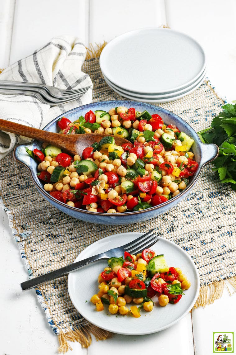 Overhead shot of a blue bowl and white plate with fork with garbanzo bean and vegetable salad on a rustic grey woven placemat with white plates and forks in the background.