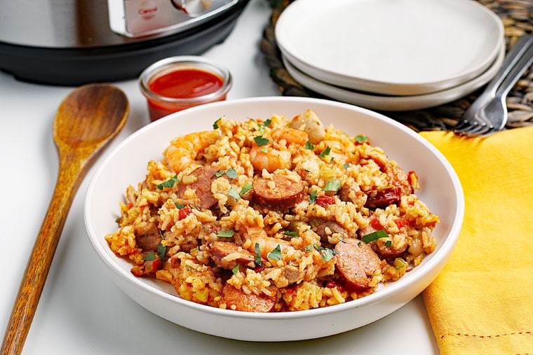 A bowl of Instant Pot Jambalaya with wooden spoon, yellow napkin, ramekin of hot sauce, pressure cooker, and white plates.