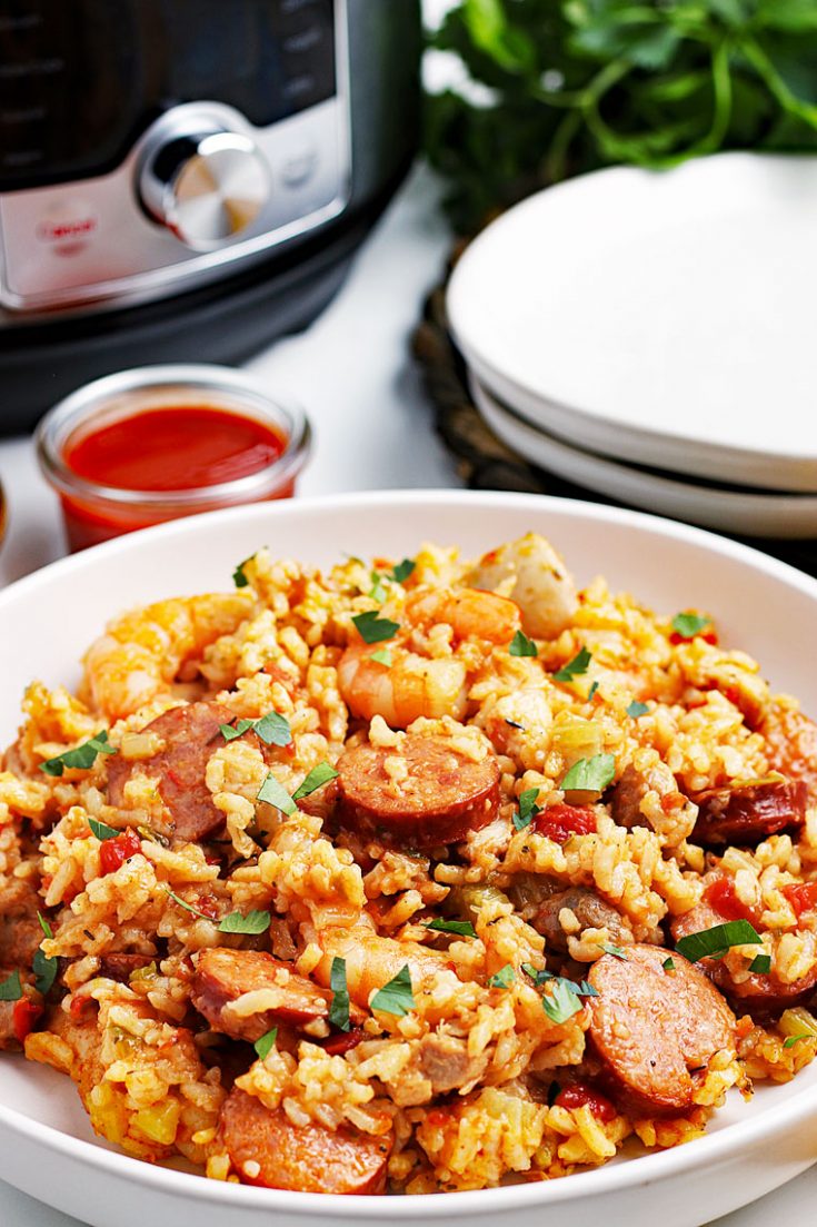 Closeup of a large bowl of jambalaya, a ramekin of hot sauce, Instant Pot pressure cooker, a stack of white plates, and a bunch of fresh parsley.