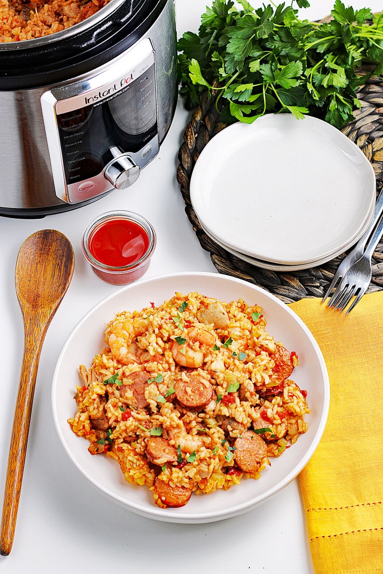 Overhead shot of a white bowl of jambalaya, a wooden spoon, a ramekin of hot sauce, Instant Pot pressure cooker, a stack of white plates, yellow napkin, and a bunch of fresh parsley.