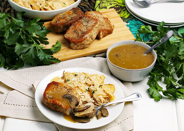 A plate of Instant Pot Pork Chops with a bowl of gravy and more chops on a wooden cutting board surrounding by herbs, napkins, and more plates.