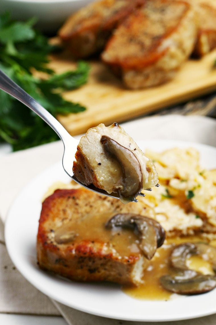 A plate and forkful of Instant Pot Pork Chops with mushrooms and potatoes with more pork chops on a cutting board in the background.
