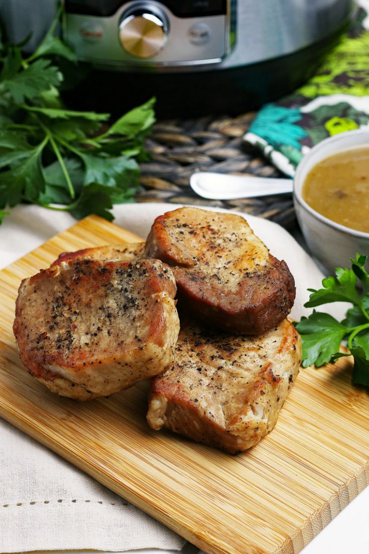 A wooden cutting board of pork chops, a bowl of gravy, and an Instant Pot in the background.